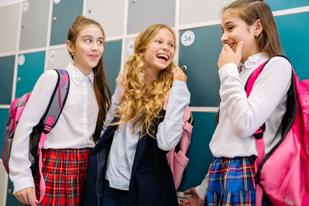 Three girls in school uniforms laughing and talking by lockers, indoors.