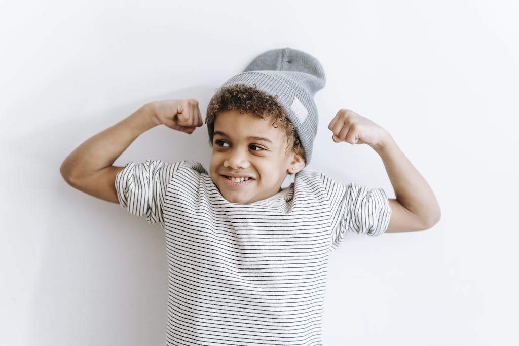 Cute boy with curly hair and beanie, flexing muscles and smiling indoors.